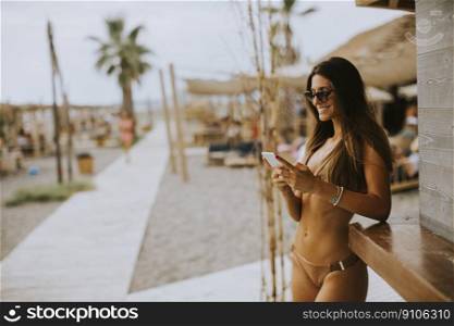 Pretty young woman in bikini standing by the surf cabin on a beach and using mobile phone at summer day