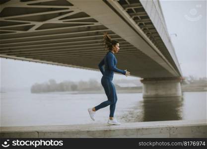 Pretty young woman in blue track suit running by the river at autumn morning