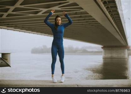 Pretty young woman in blue track suit stretching before workout by the river at autumn morning