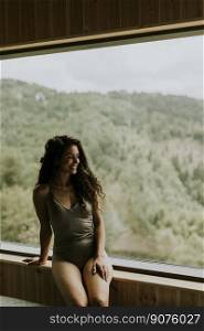 Pretty young woman in wellness spa center sitting by the huge wide window enjoying the green forest view