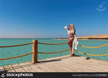 Pretty young woman in white pareo standing on pier near sea.. Pretty young woman in white pareo standing on pier near sea