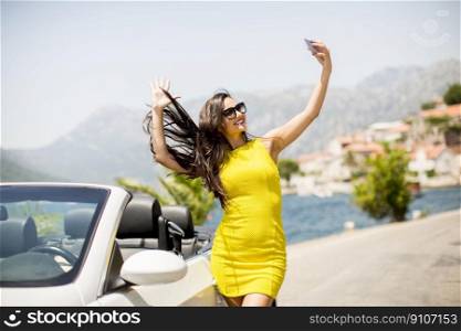 Pretty young woman in yellow dress taking selfie with mobile phone by white cabriolet car