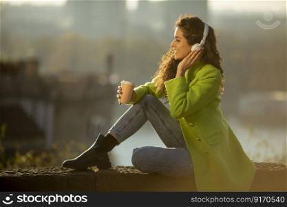 Pretty young woman listening music with smartphone and takeaway coffee by the river at sunny autumn day