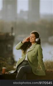 Pretty young woman listening music with smartphone and takeaway coffee by the river at sunny autumn day
