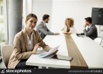 Pretty young woman sitting by the table with digital tablet and paper chart in modern office in front of her team
