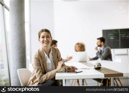 Pretty young woman sitting by the table with digital tablet and paper chart in modern office in front of her team