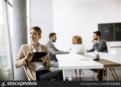 Pretty young woman sitting by the table with digital tablet in modern office in front of her team