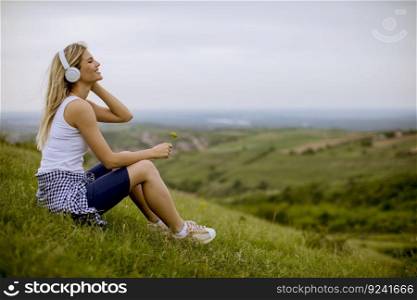 Pretty young woman sitting in nature, holding a small flower and listening to music on headphones