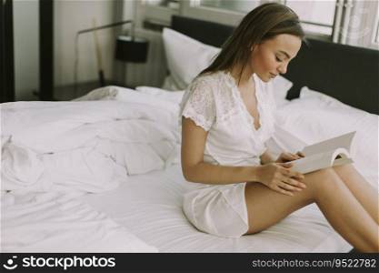 Pretty young woman sitting on a bed and reading a book