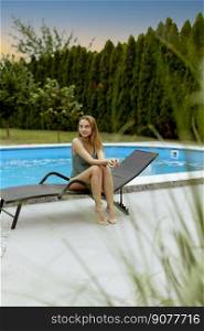 Pretty young woman sitting on a deck char by the swimming pool in the house backyard