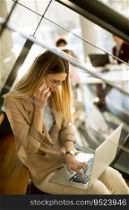 Pretty young woman sitting on the stairs with laptop and using mobile phone in modern office in front of her team