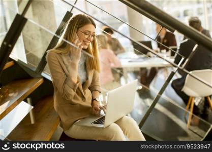Pretty young woman sitting on the stairs with laptop and using mobile phone in modern office in front of her team