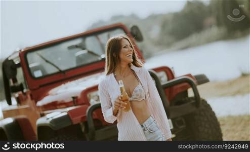 Pretty young woman standing by a red car, drinks refreshment and enjoys it