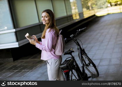 Pretty young woman standing by an electric bicycle and using digital tablet in urban environment