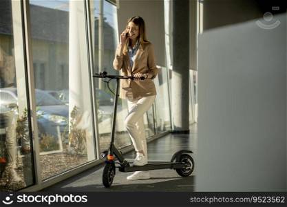 Pretty young woman standing by the office window with electric scooter and using mobile phone