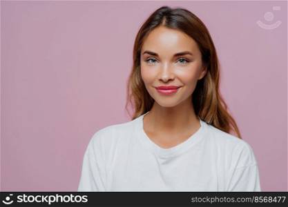 Pretty young woman uses cosmetics for looking young and beautiful, has long hair, looks confidently at camera with happy tender smile, wears casual outfit, poses over purple wall, blank space on right