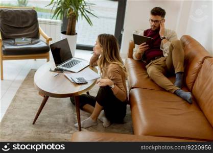 Pretty young woman using laptop and young man using digital tablet while sitting on sofa at home