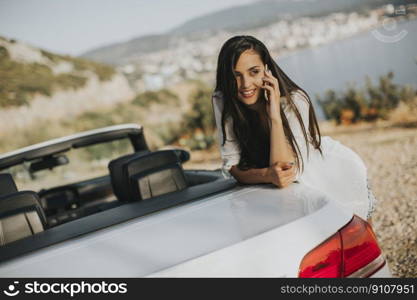 Pretty young woman using mobile phone and standing by cabriolet car on a hot summer day