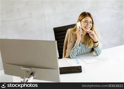 Pretty young woman using mobile phone while sitting by the desk in the office