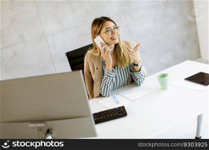Pretty young woman using mobile phone while sitting by the desk in the office