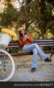 Pretty young woman using phone in autumn park sitting on bench by the bicycle
