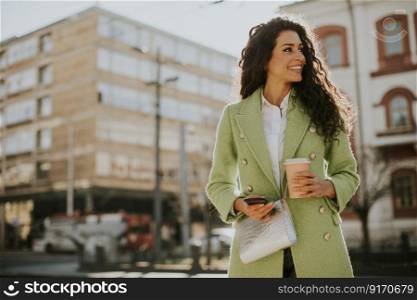 Pretty young woman using smartphone on the street and holding takeaway coffee