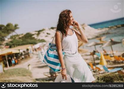 Pretty young woman with a bag using mobile phone on the beach at sunny summer day