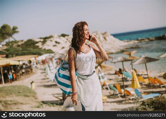 Pretty young woman with a bag using mobile phone on the beach at sunny summer day