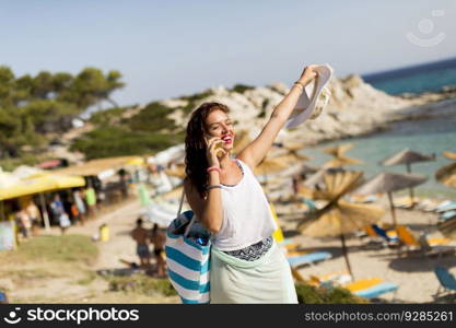 Pretty young woman with beach bag talking on mobile phone by the sea at summer