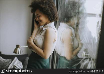 Pretty young woman with curly hair using mobile phone by the window at home