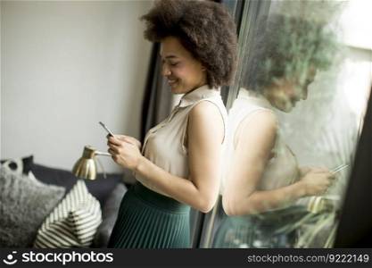 Pretty young woman with curly hair using mobile phone by the window at home