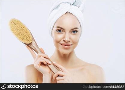 Pretty young woman with healthy skin, takes care of personal hygiene, holds bath brush, wrapped towel on head, stands shirtless indoor, white background. Women, skin care, freshness concept.
