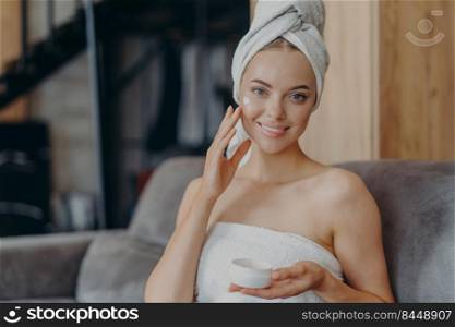 Pretty young woman with healthy smooth skin applies face cream, wears wrapped towel on head after taking shower, poses on comfortable sofa. Hygiene procedure, cosmetology and beauty concept.