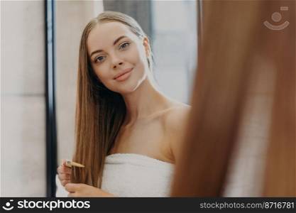 Pretty young woman with minimal makeup, full lips looks at herself in mirror, combs long straight hair, wrapped in bath towel, cares about herself, wants to have fabulous look. Hair care concept
