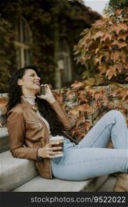 Pretty young woman with mobile phone drink coffee to go at the outdoor stairs on autumn day