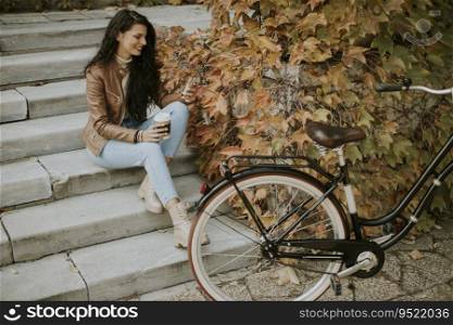 Pretty young woman with mobile phone drink coffee to go at the stairs by the bicycle on autumn day