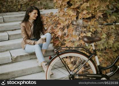 Pretty young woman with mobile phone drink coffee to go at the stairs by the bicycle on autumn day