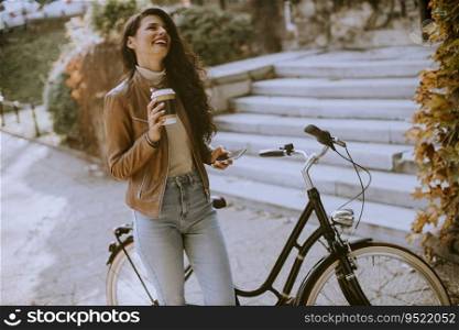 Pretty young woman with mobile phone drink coffee to go by the bicycle on autumn day