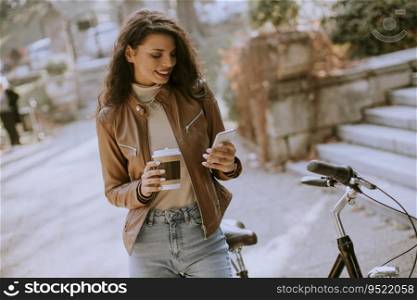 Pretty young woman with mobile phone drink coffee to go by the bicycle on autumn day
