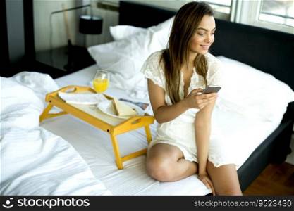 Pretty young woman with mobile phone sitting on the bed in the room