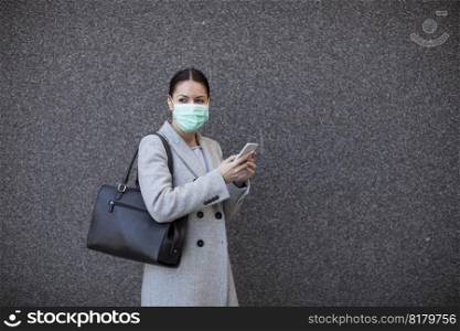 Pretty young woman with protective facial mask on the street using mobi≤pho≠