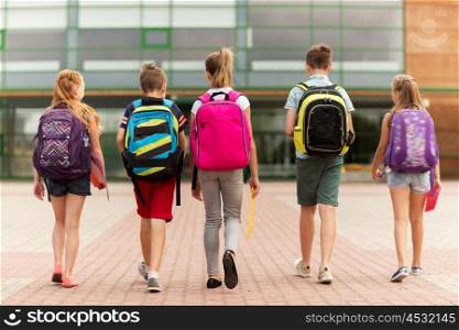 primary education, friendship, childhood and people concept - group of happy elementary school students with backpacks walking outdoors from back