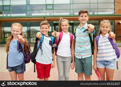 primary education, friendship, childhood and people concept - group of happy elementary school students with backpacks pointing finger on you outdoors