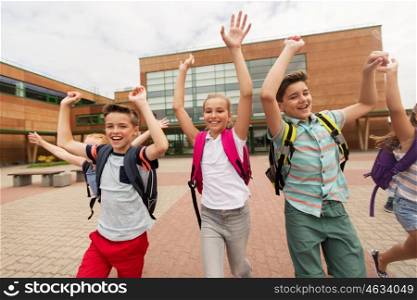 primary education, friendship, childhood and people concept - group of happy elementary school students with backpacks running and waving hands outdoors