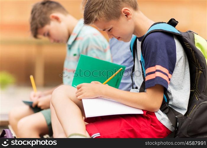 primary education, friendship, childhood, communication and people concept - elementary school student boys with backpacks writing to notebooks and sitting on bench outdoors