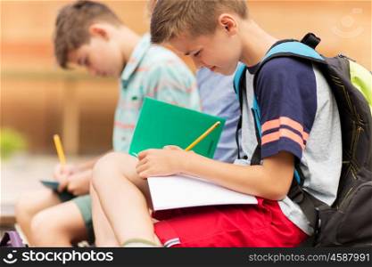 primary education, friendship, childhood, communication and people concept - elementary school student boys with backpacks writing to notebooks and sitting on bench outdoors
