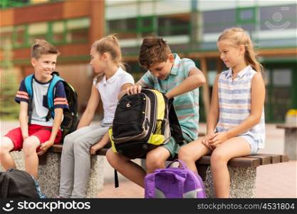 primary education, friendship, childhood, communication and people concept - group of happy elementary school students with backpacks sitting on bench outdoors