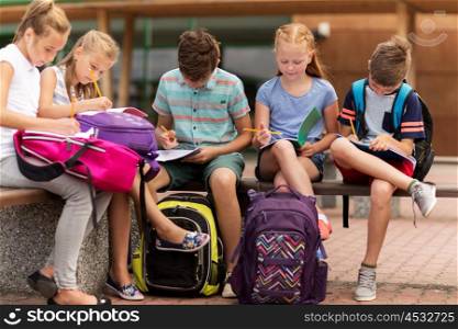 primary education, friendship, childhood, communication and people concept - group of happy elementary school students with backpacks and notebooks sitting on bench and doing homework outdoors