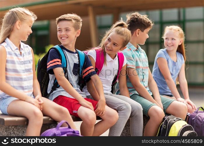 primary education, friendship, childhood, communication and people concept - group of happy elementary school students with backpacks sitting on bench and talking outdoors