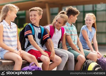 primary education, friendship, childhood, communication and people concept - group of happy elementary school students with backpacks sitting on bench and talking outdoors